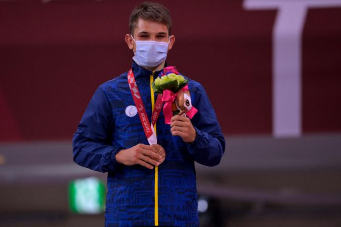 A male athlete wearing a face mask poses for a photograph after receiving a bronze medal. He is holding a bouquet with his left hand.