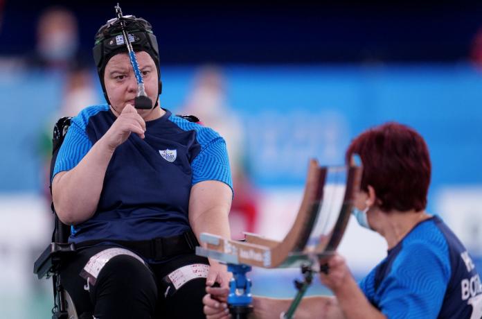 Anna Ntenta, a female boccia player from Greece, is looking at her boccia ramp during competition while her sport assistant adjusts it. Ntenta is wearing a head gear with a pointer.