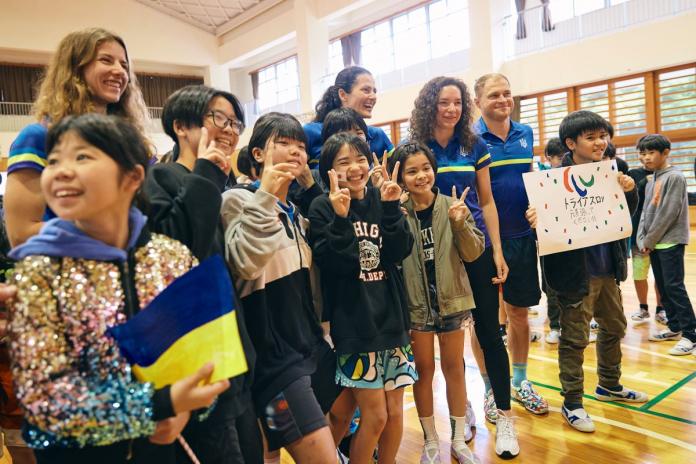 Para triathletes from Ukraine are posing for a photo with Japanese pupils. A boy is holding a sign with the Three Agitos.
