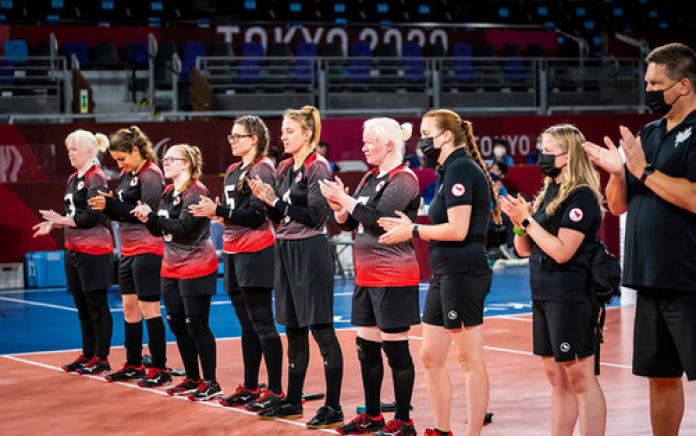 Eight female goalball players and a male staff clap their hands while lining up on court