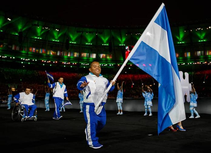 A man of short stature smiling and waving at the camera while holding a flag 