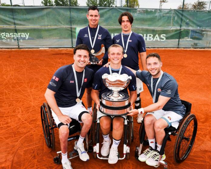 Five male athletes pose for a photograph. A young wheelchair tennis player in the centre is holding a silver trophy.