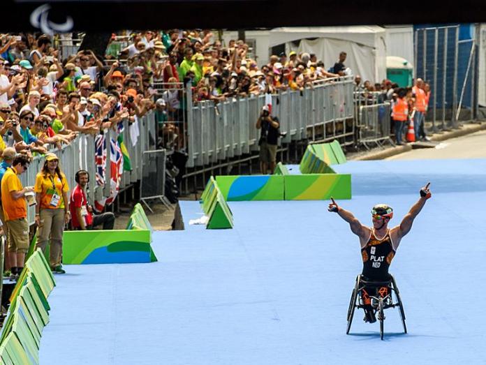 A Para triathlete competing in a racing wheelchair raises his arms in front of a crowd