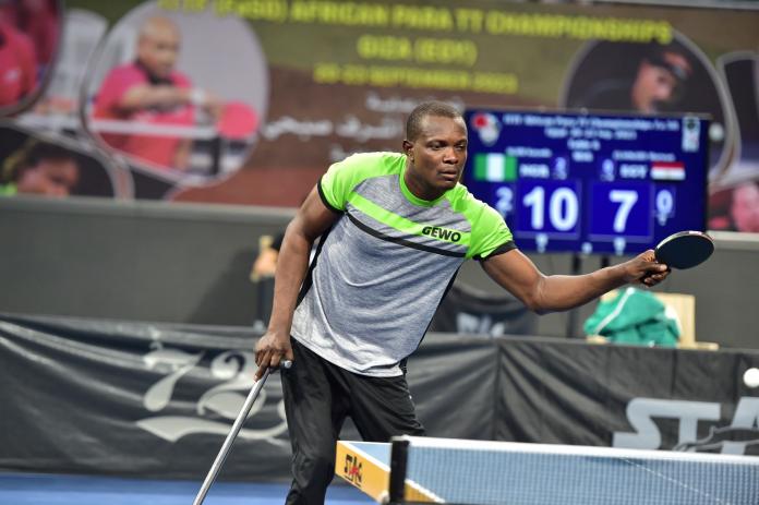 A male Para table tennis player in action. He is holding a cane with his right hand and a table tennis paddle with his left.