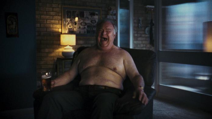 A naked man sits on a couch drinking beer