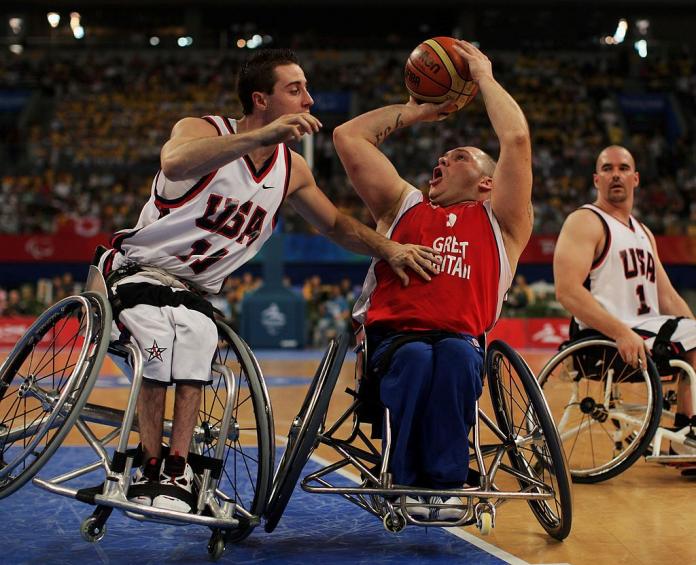 Dos jugadores masculinos de baloncesto en silla de ruedas están en acción. Un jugador está intentando un lanzamiento, mientras que otro jugador se inclina para bloquear.