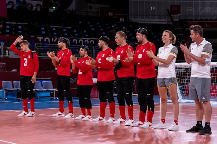 Seis atletas de goalball masculino y dos miembros del personal se paran en línea en la cancha y aplauden