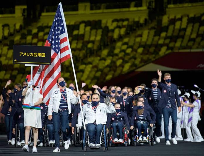 A group of US Para athletes led by a man using a wheelchair and holding the American flag 