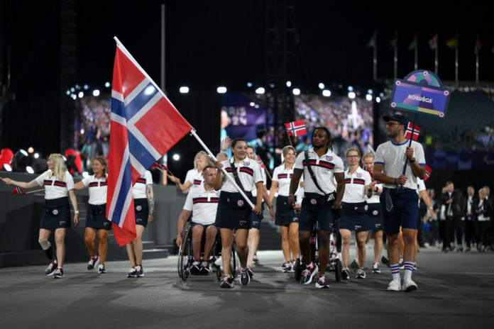 A crowd of Norwegian athletes enter a ceremony in matching uniforms and waving Norwegian flags