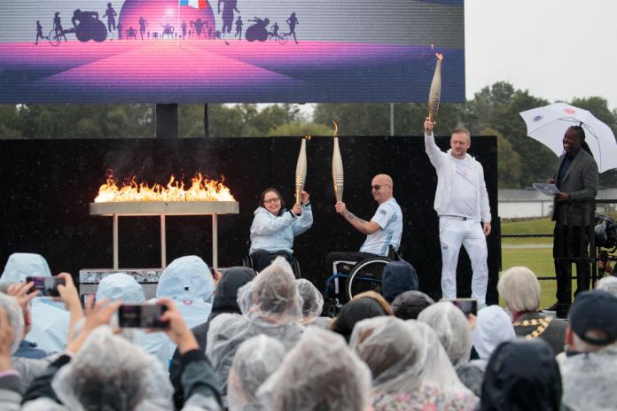 two Paralympians in wheelchairs and a man standing holding up Paralympic torches