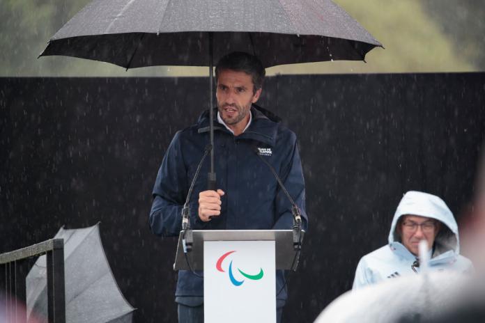 Paris 2024's Tony Estanguet holding an umbrella and speaking at a podium 