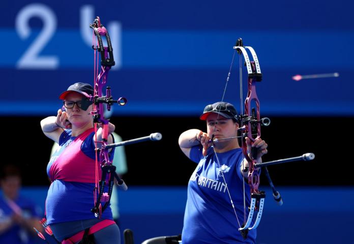 Jodie on the left in pink and blue with an obvious baby bump and Phoebe Paterson Pine on the right in blue Great Britain t-shirt both raise their bows to eye level to line up a shot