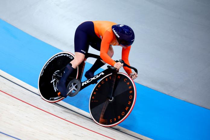 Cyclist in orange top, blue bottoms and blue helmet leans forward on her bike in velodrome