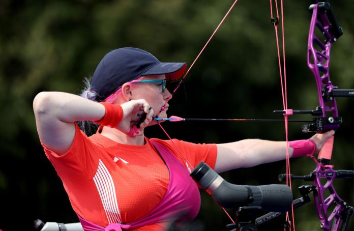 A woman in red and a cap with pink hair focuses her archery bow
