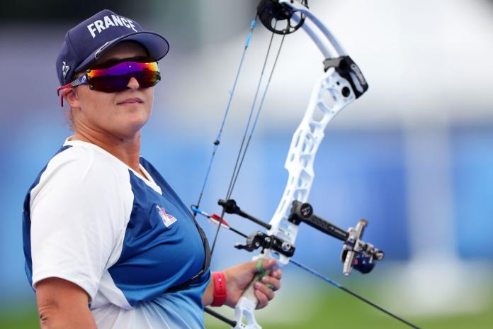 Woman in France cap and rainbow sunglasses turns to face the camera holding an archery bow