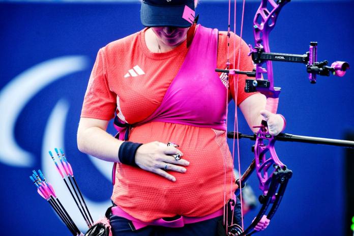 Woman in blue hat, red t-shirt and pink protector holding archery bow looks down at her baby bump holding a hand on it
