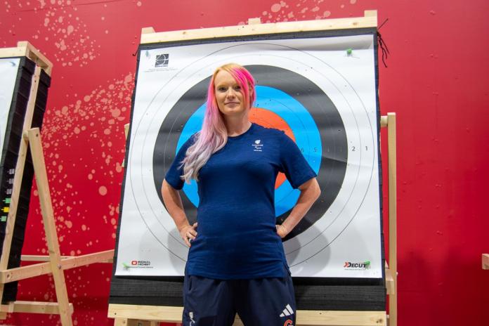 A woman in blue with long pink hair stands with hands on hips in front of Archery target with visible baby bump