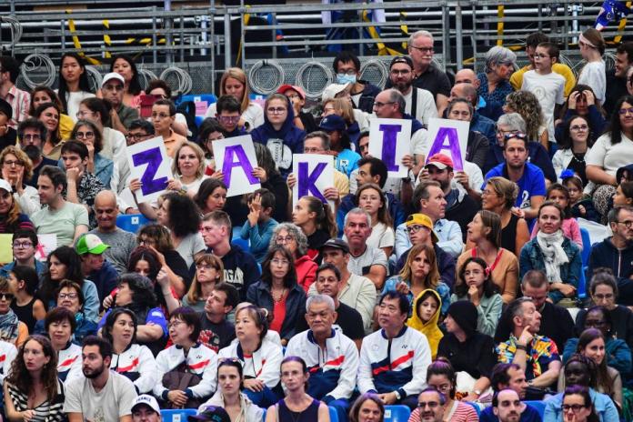 A wide shot of the crowd at the Grand Palais. Five spectators are holding a poster that says Zakia