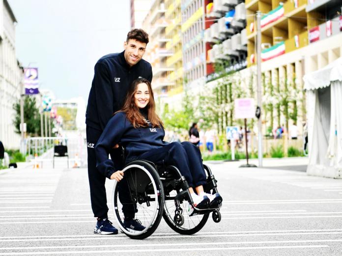 A man pushes a woman in a wheelchair, they're both in dark blue and smiling at the camera