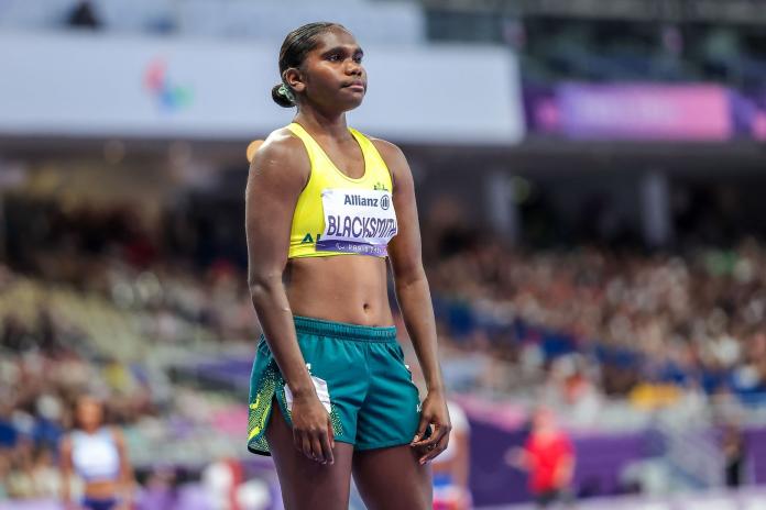 An Australian female athlete with a yellow vest and green shorts prepares to start her race