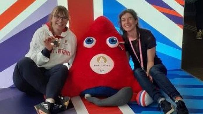Para athlete Anna Nicholson holds up a bronze medal sitting next to a friend