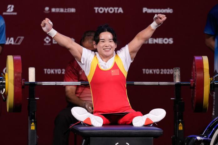 A woman on a powerlifting bench sits up and raises her arms in the air in celebration