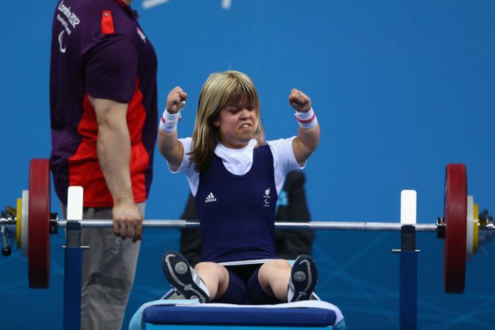 A woman on a powerlifting bench sits up and raises her arms in the air in celebration
