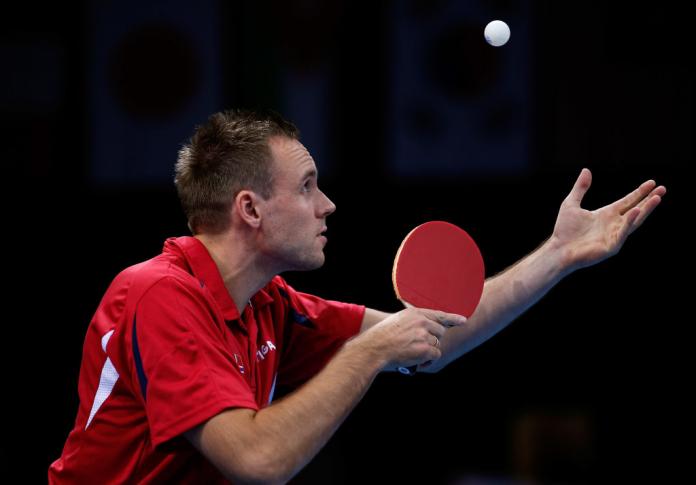 A man holds a table tennis paddle in one hand looks up at table tennis ball in the air.