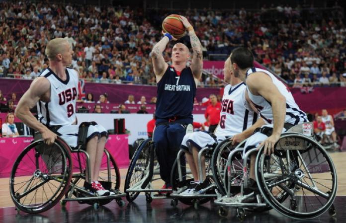 A man in a wheelchair and a blue uniform holds a basketball above his heads and prepares to shoot while surrounded by three other wheelchair basketball players in white uniforms