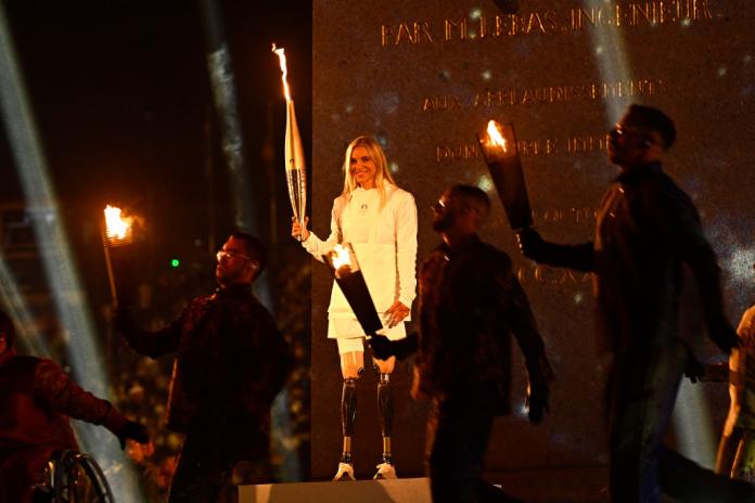 A woman with prosthetic legs and wearing all white holds up the Paralympic torch in front of a large stone monument