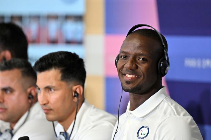 A man wearing a white polo shirt and headphones smiles at the camera