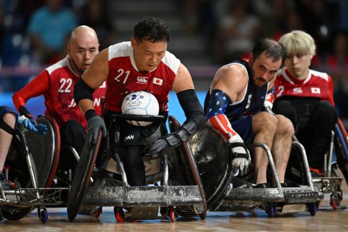 Four men in wheelchairs play wheelchair rugby. One holds the ball in his lap and the others follow him.