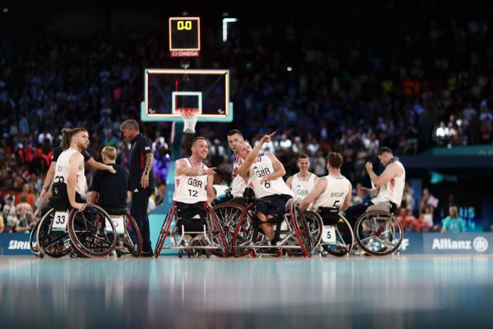 Seven men in white uniforms and wheelchairs gather on a basketball court. Two players point at something not visible in the picture.