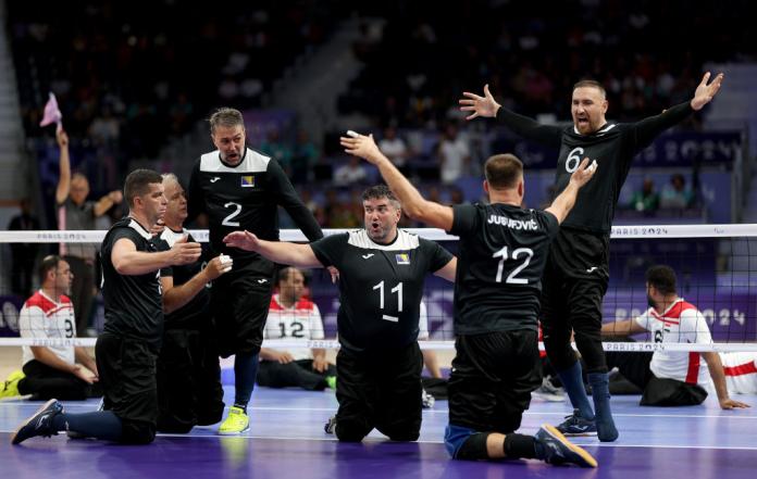 Six men in black uniforms celebrate a point in sitting volleyball, raising their arms in the air and cheering