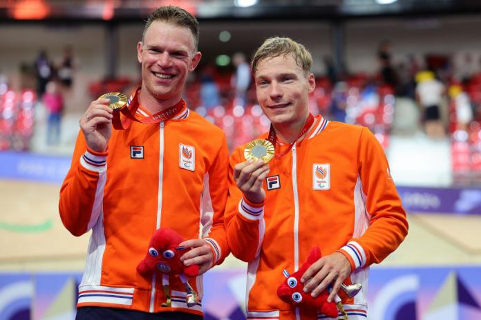 Two men in orange Team Netherlands jackets smile and hold up gold medals