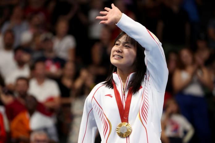 Woman in white and red clothing with a gold medal around her neck raises her hand and waves with a smile