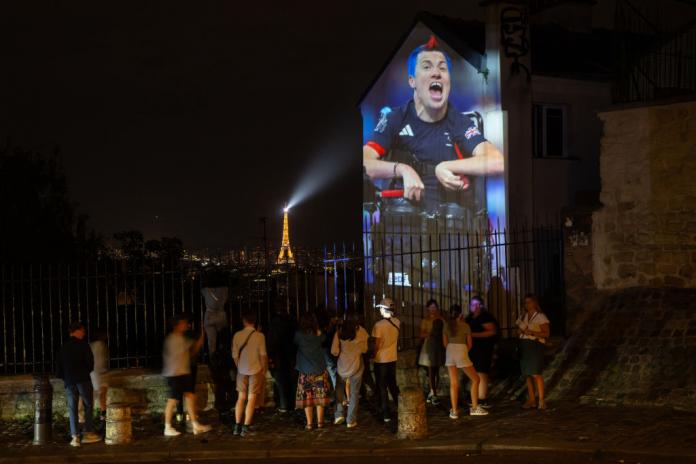 A photo of a man in a wheelchair is projected onto the side of a building. A crowd of people stand by the building. The Eiffel Tower is in the distance. It is night.