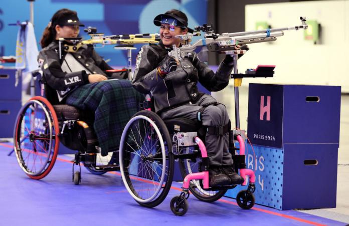 A woman in a wheelchair holds up her shooting Para sport rifle and smiles