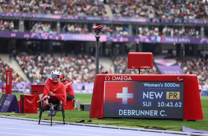 A woman in a racing wheelchair smiles for the camera next to an LED screen that displays her race time
