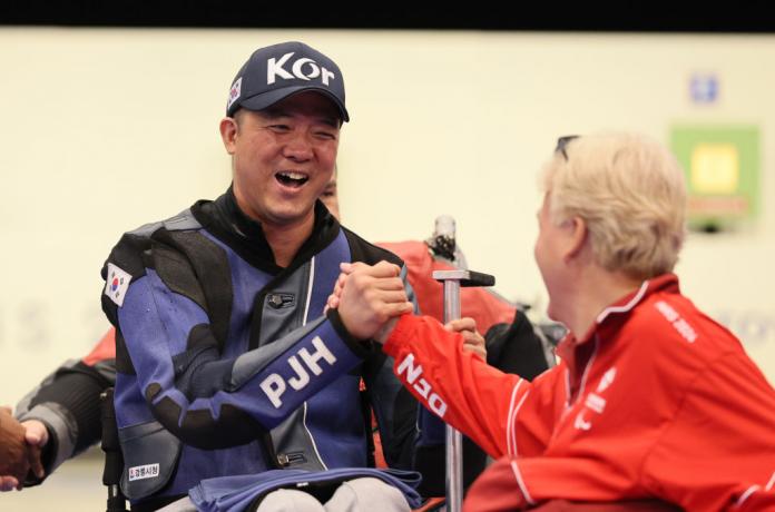 A man in a baseball hat and shooting Para sport jacket and smiles and grabs the hand of a man in a red jacket in celebration