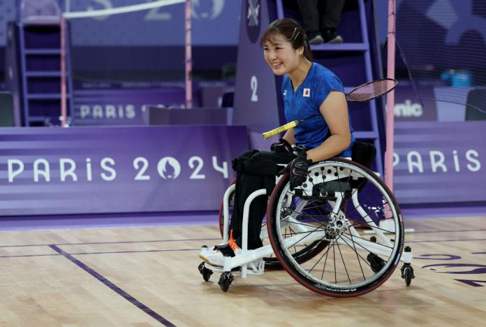 A woman in a wheelchair holds a badminton racquet under her arm and smiles