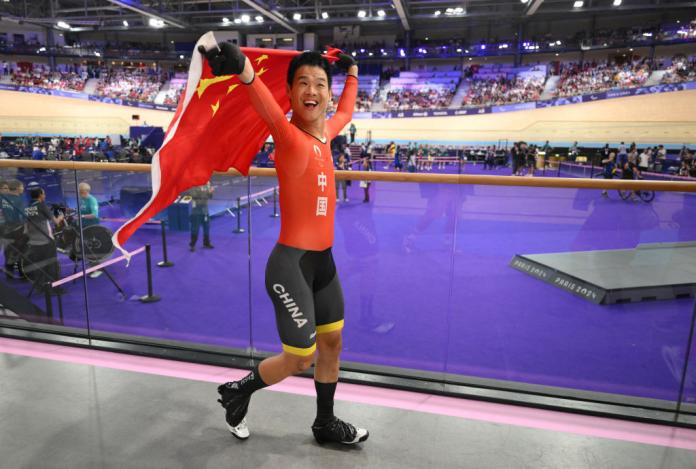 A man in a Team China uniform and cycling shoes walks on a walkway inside a cycling velodrome, smiling and holding up a Chinese flag