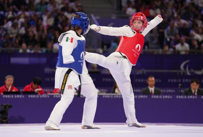 Two women compete in Para taekwondo, one in a red helmet and vest, the other in blue. The athlete in red kicks the athlete in blue.