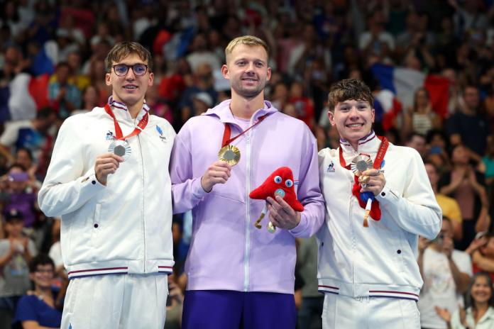 Three men stand on a podium and hold up medals, with a big crowd behind them