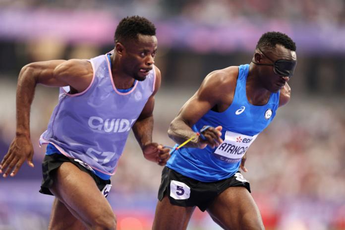 Two men run, one wears an eye covering, the other has a 'guide' bib.