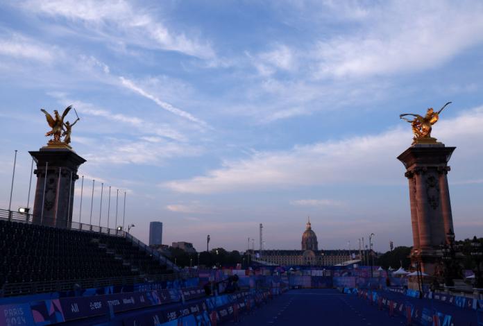 A view of the finish line for a Para triathlon event. There are spectator stands and tall towers with golden figurines. A large historical building is visible in the background.