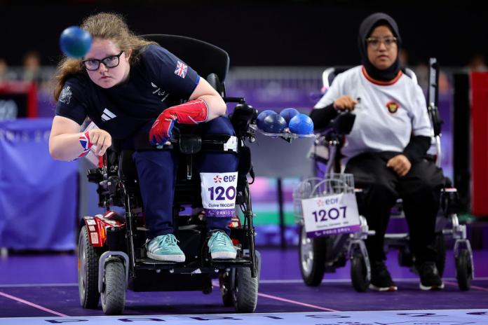 Two women in wheelchairs play boccia. One woman has just thrown a blue ball.