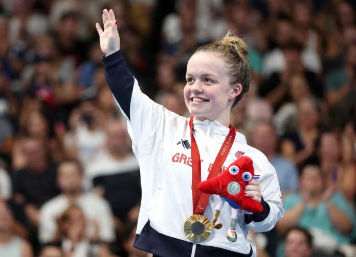 A woman wearing a gold medal and holding a red stuffed mascot waves