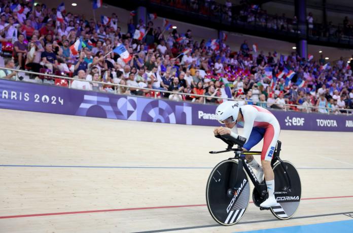 A woman in a Team France uniforms and rides a bike inside a velodrome as a large crowd waves French flags 