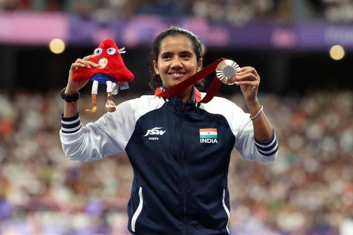 A woman in a Team India jacket holds up the Paris 2024 mascot in one hand and a bronze medal in the other